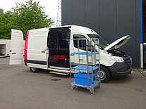 Weißer Transporter mit offener Seitentür und Motorhaube, blauer Wagen in der Nähe.